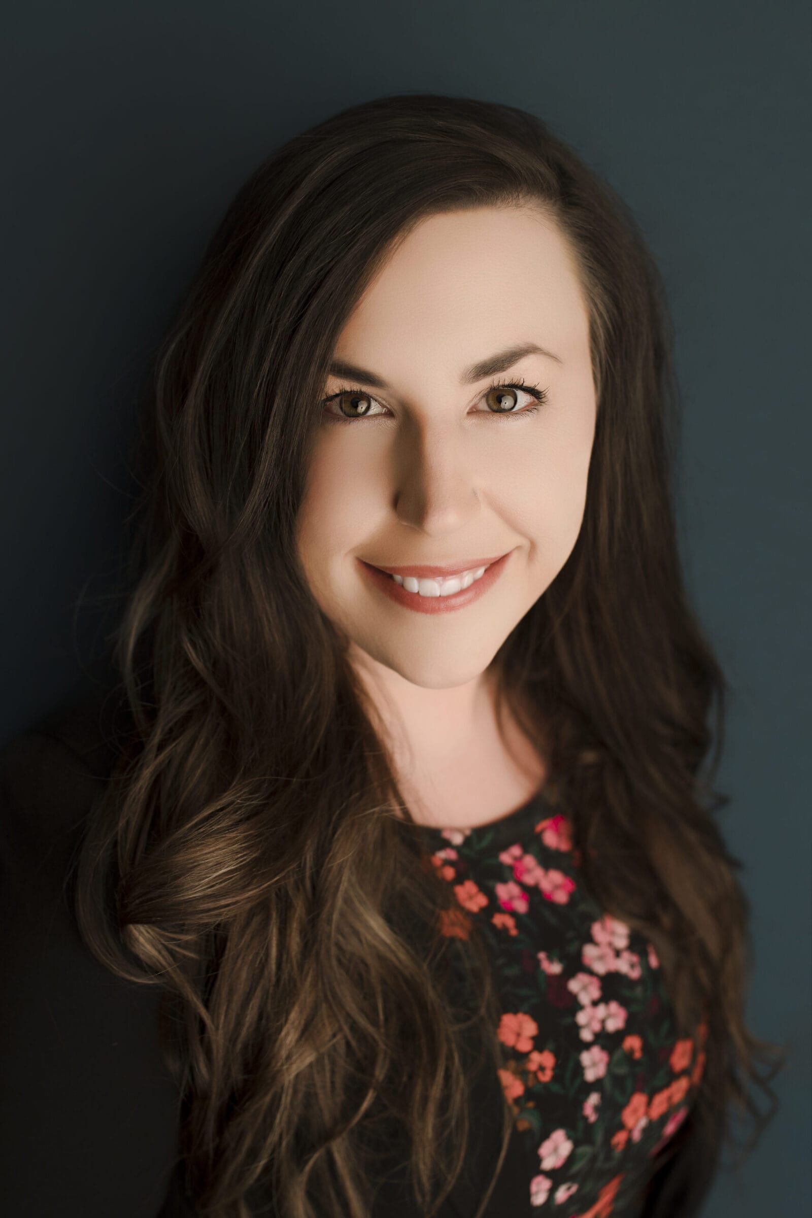 Smiling woman with long, wavy hair, wearing a floral top against a dark blue background.