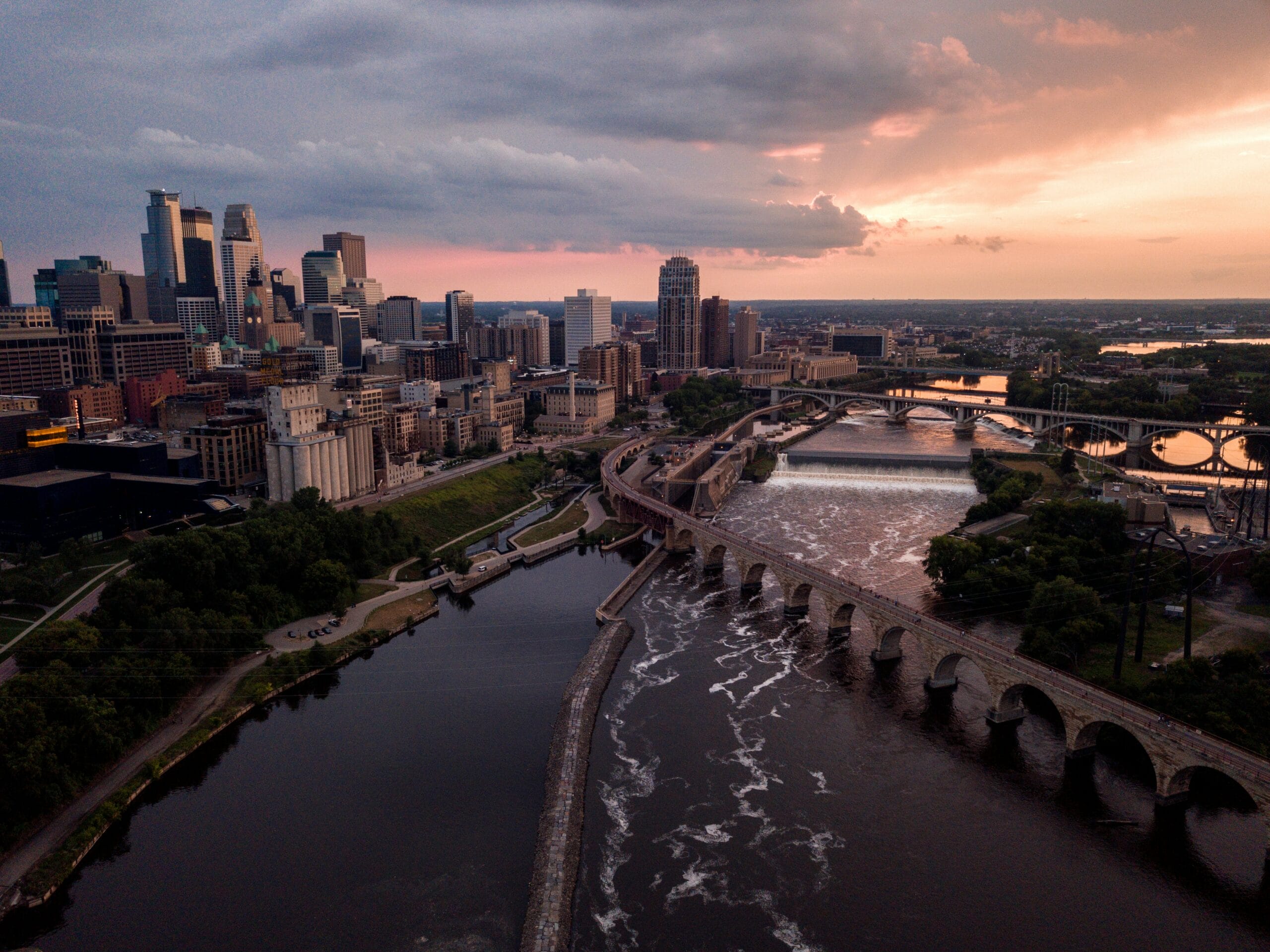 Sunset over Minneapolis skyline, featuring bridges and the Mississippi River with flowing water and lush greenery.