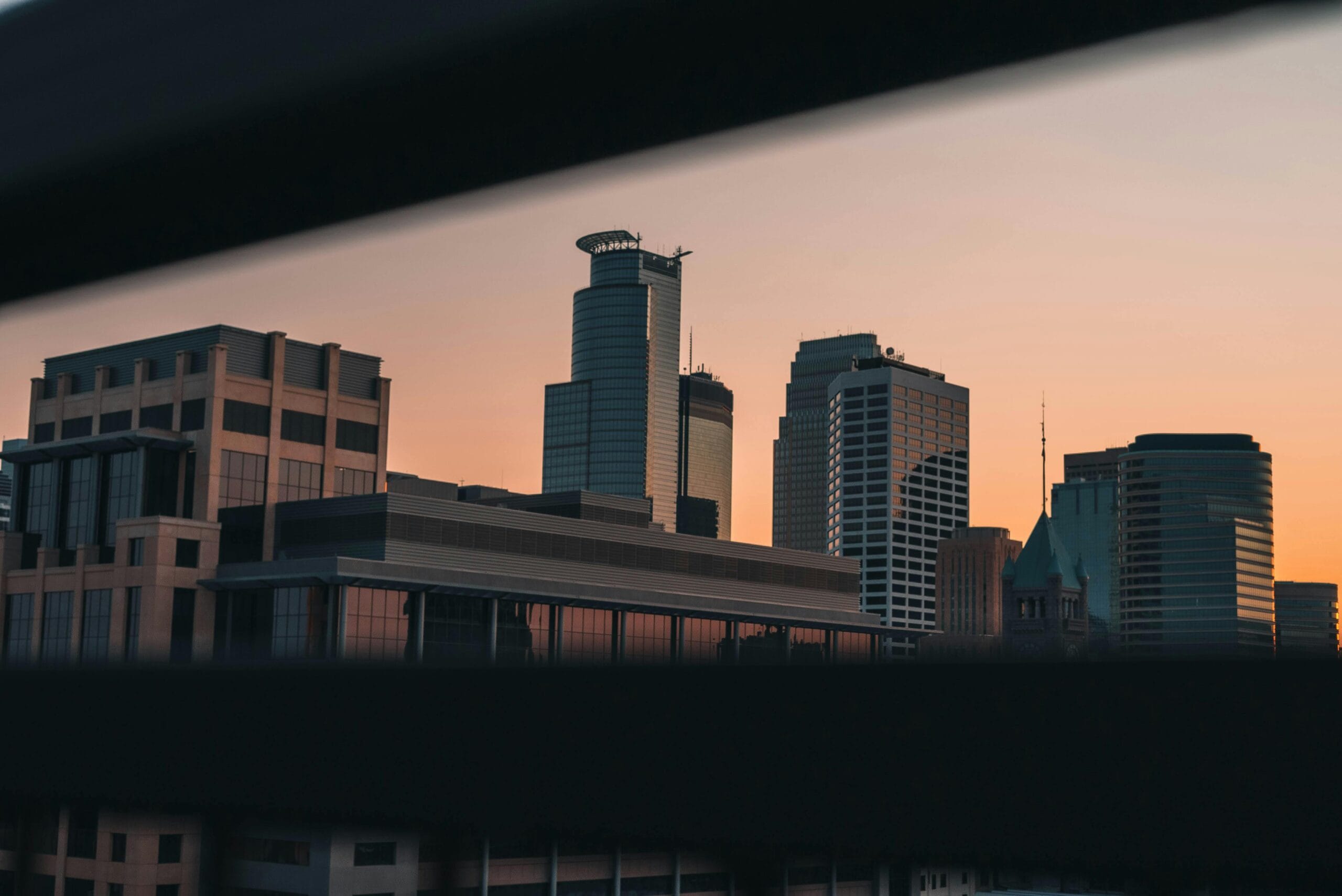 City skyline at sunset, featuring modern skyscrapers and a historic building, framed by a dark foreground.