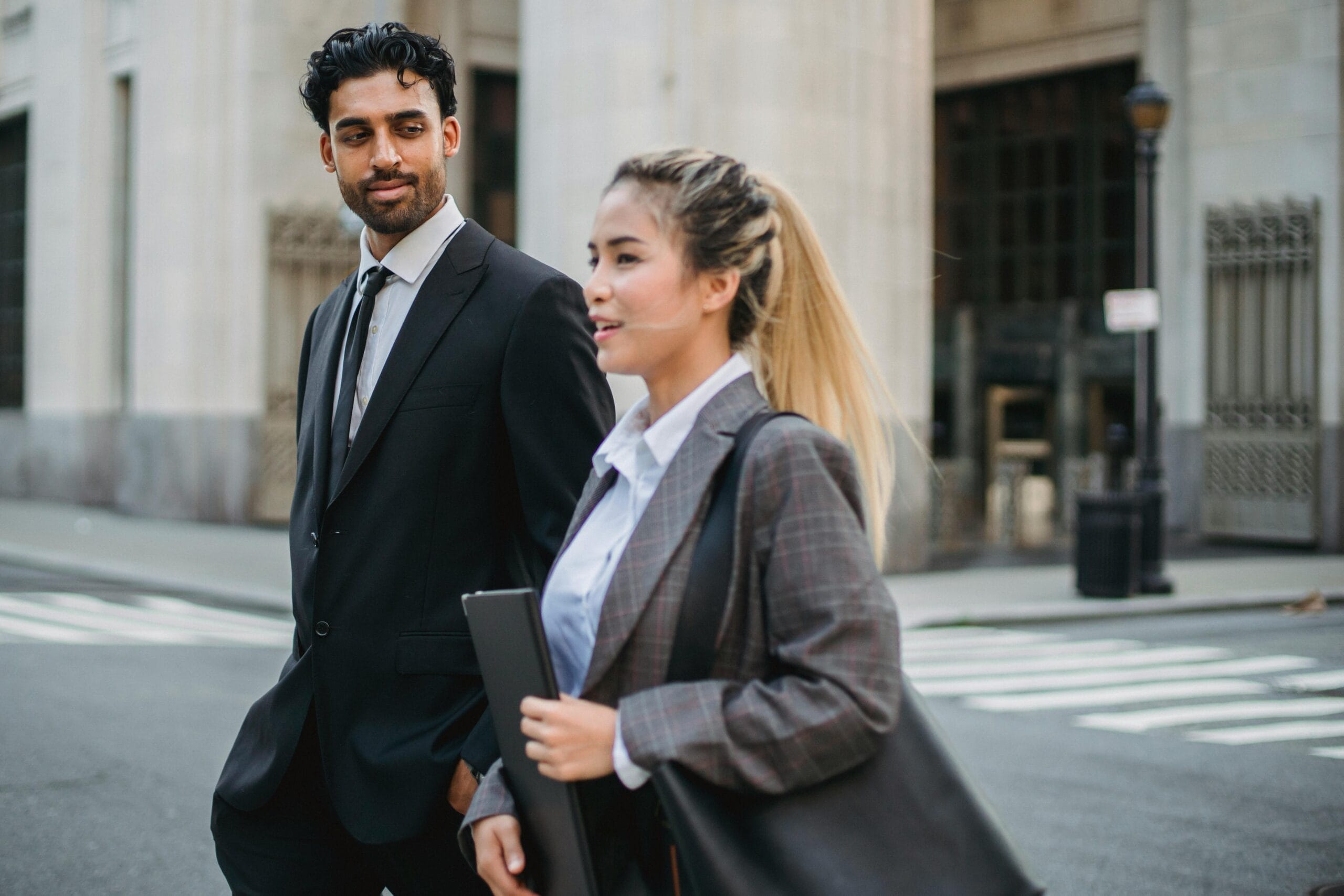 A man in a suit and a woman in a plaid blazer walk together on a city street, engaged in conversation.