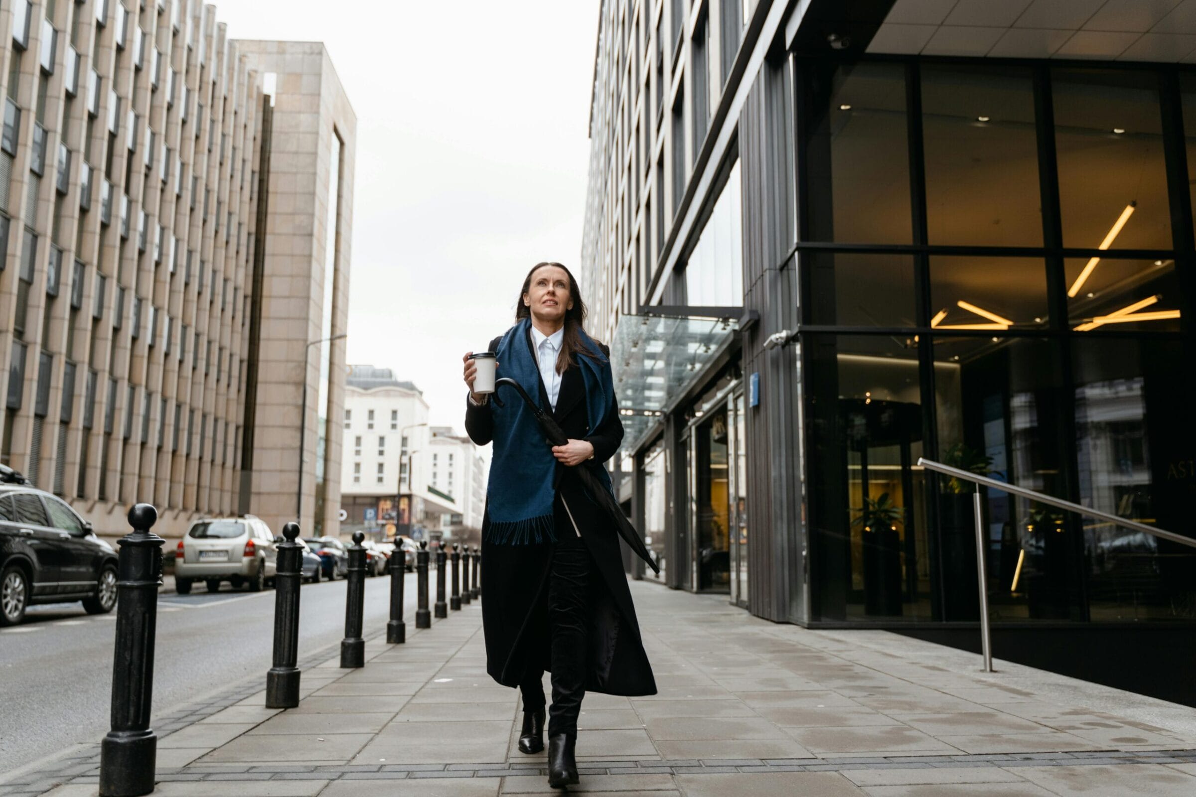 A woman in a long coat and scarf walks down a city street, holding a coffee cup, with modern buildings in the background.