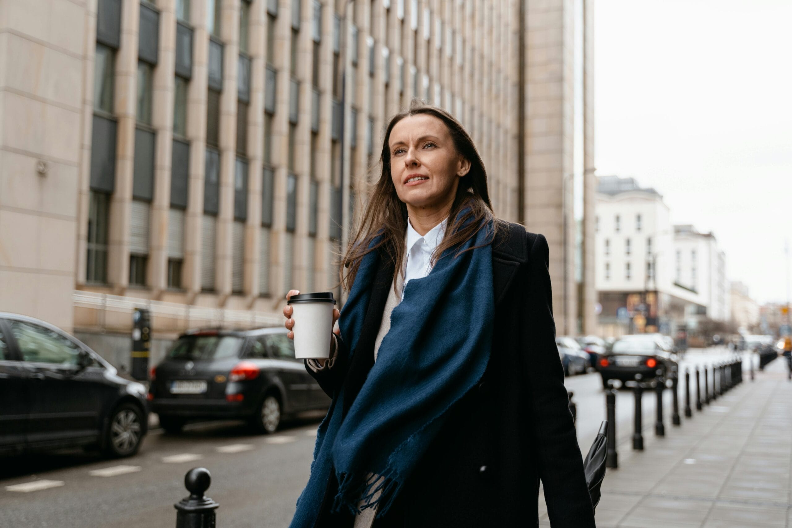 Woman in a black coat and blue scarf walks on a city street, holding a coffee cup, with cars and buildings in the background.