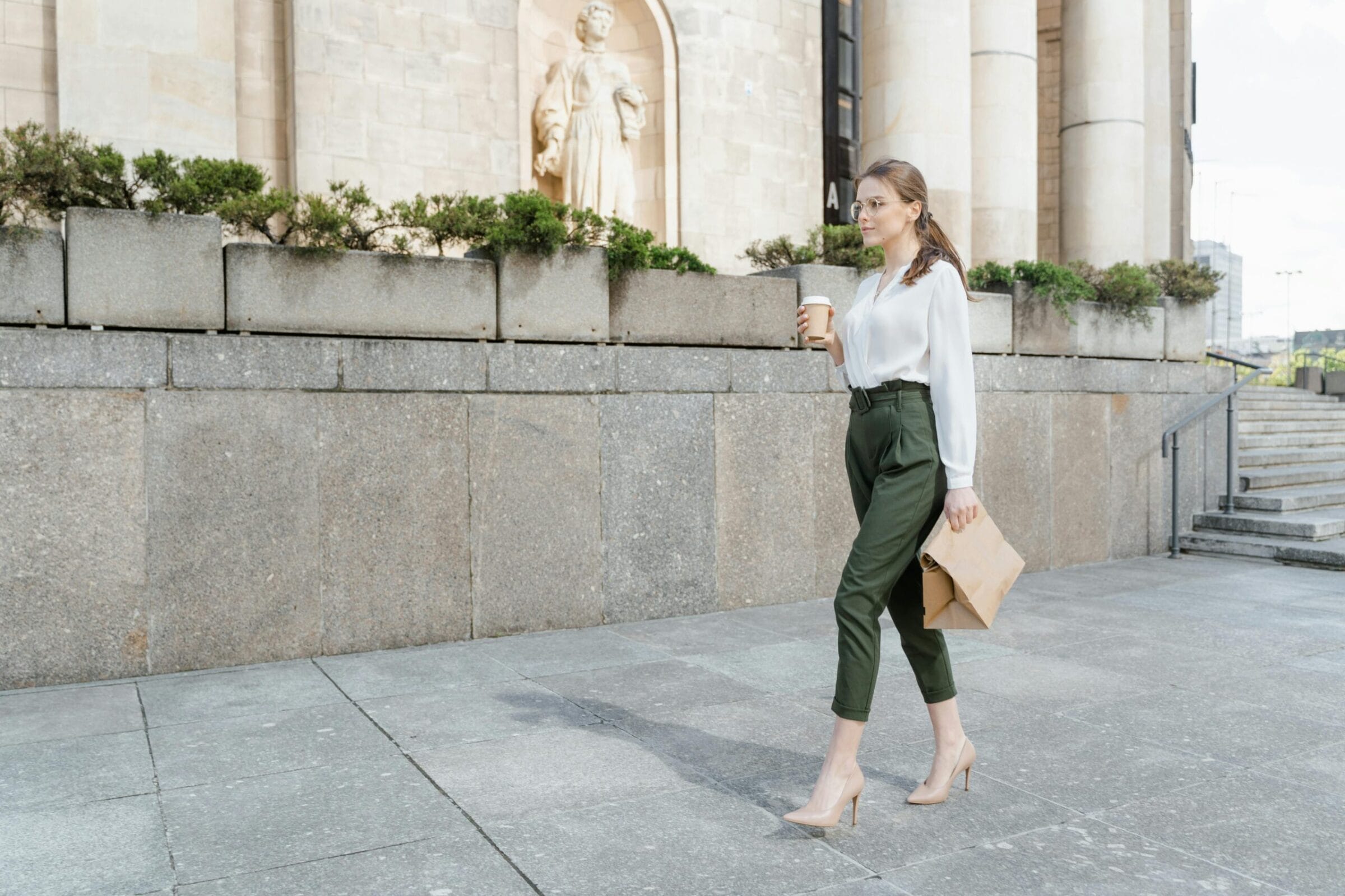 A woman in a white blouse and green trousers walks with a coffee cup and a folder, near a stone wall and greenery.