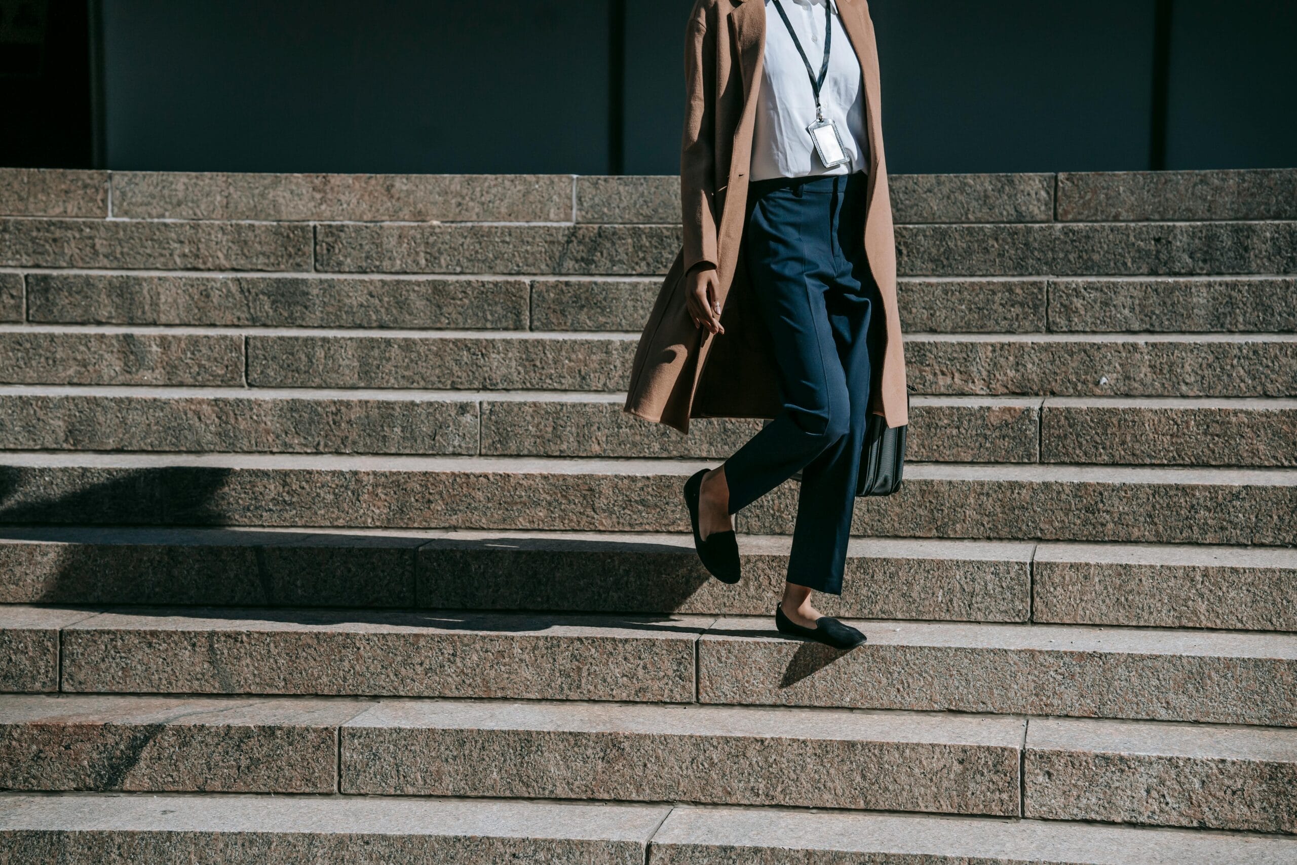 A person in a long coat and smart attire descends stone steps, holding a bag and wearing black flats.