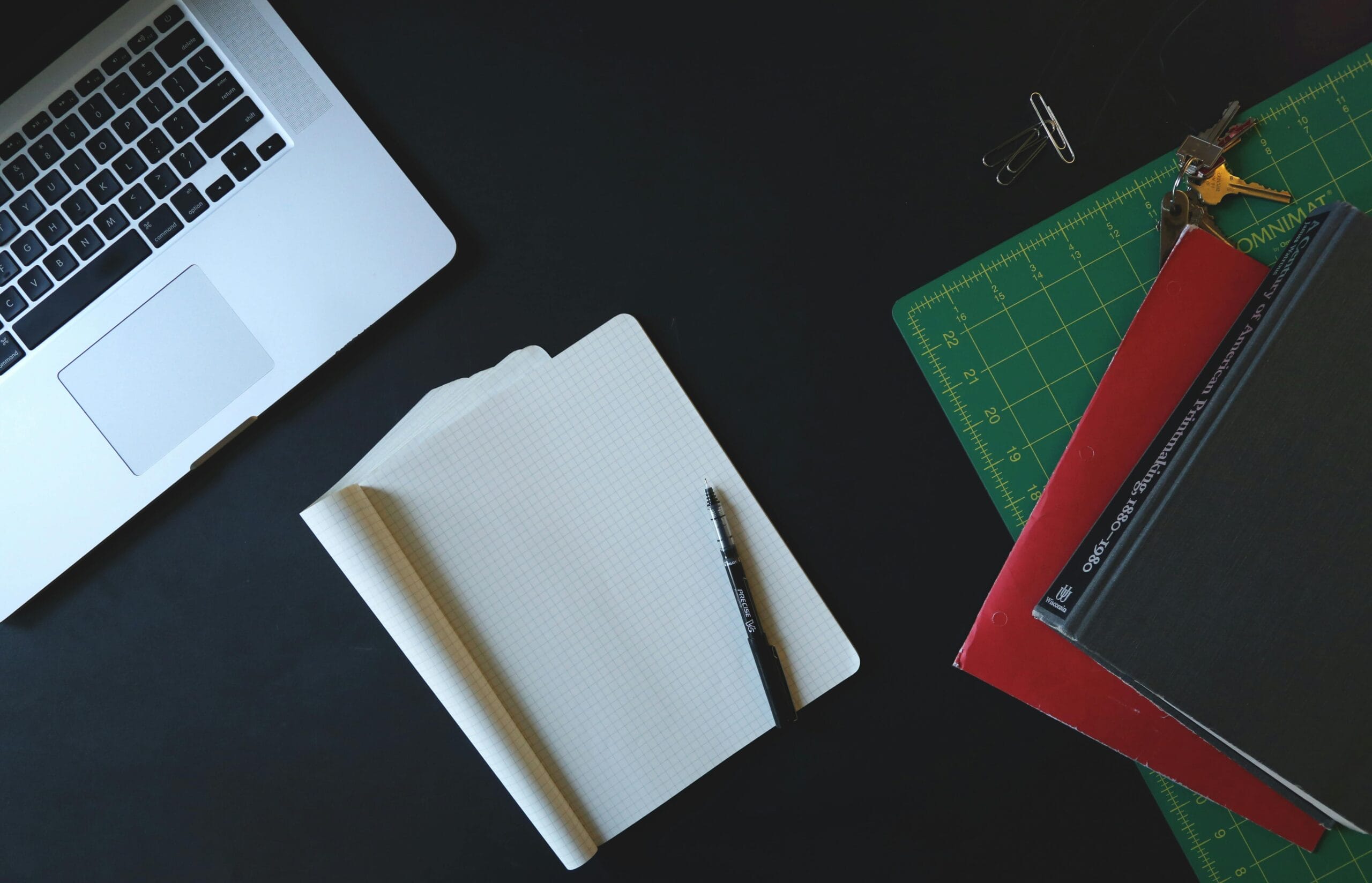 Laptop, blank notebook with a pen, and stacked folders on a dark desk, with a cutting mat and paper clips nearby.