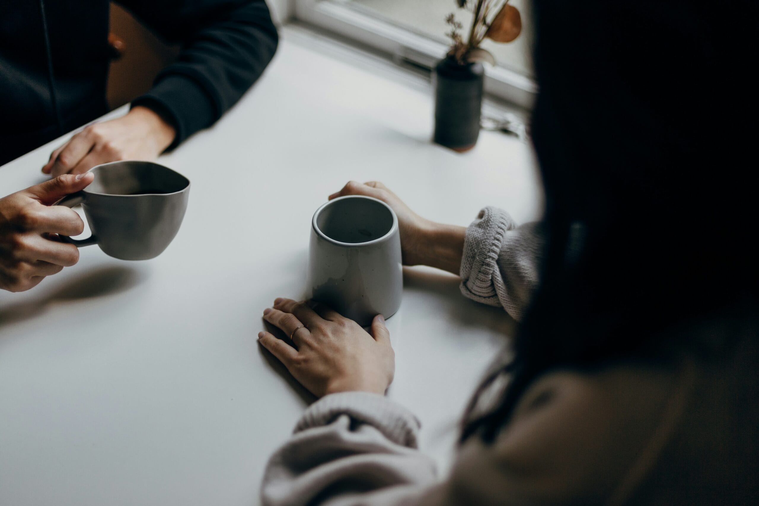 Two hands holding mugs on a table, with a small potted plant in the background and soft natural light coming through a window.