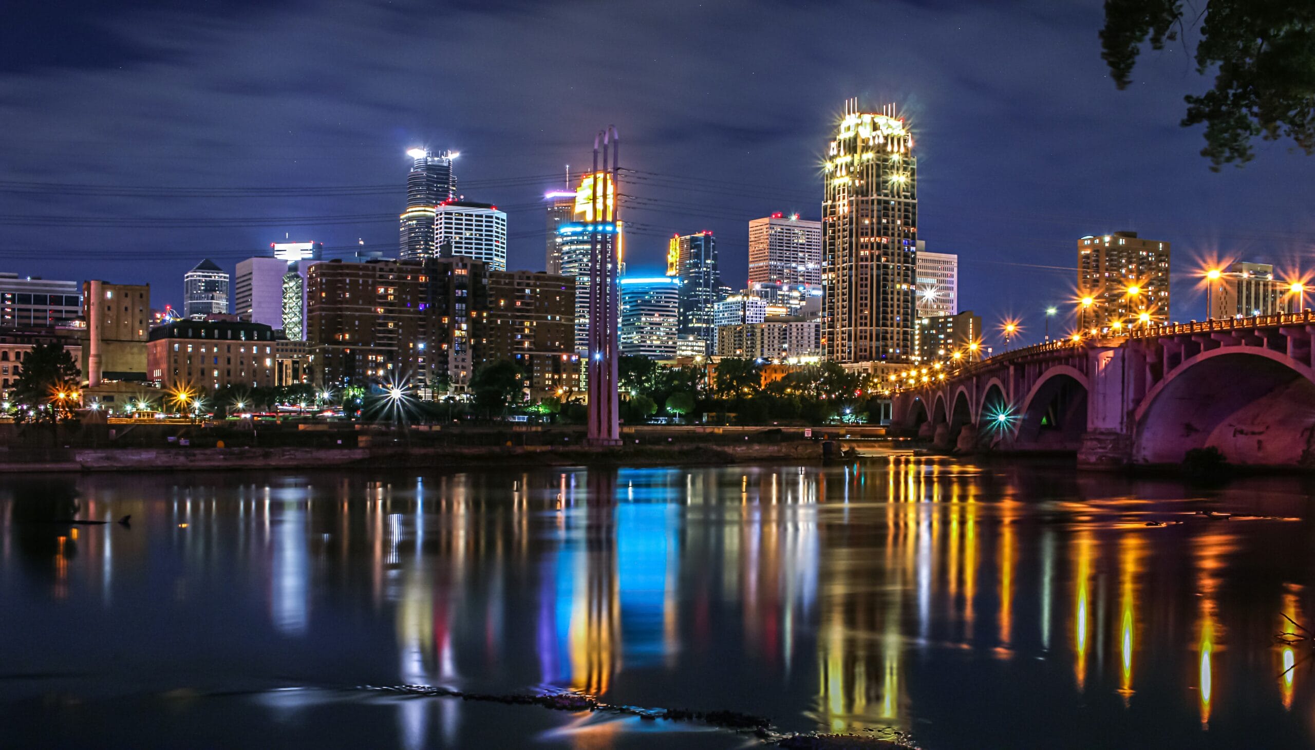 City skyline illuminated at night, reflecting in calm water, with a bridge arching across the foreground.